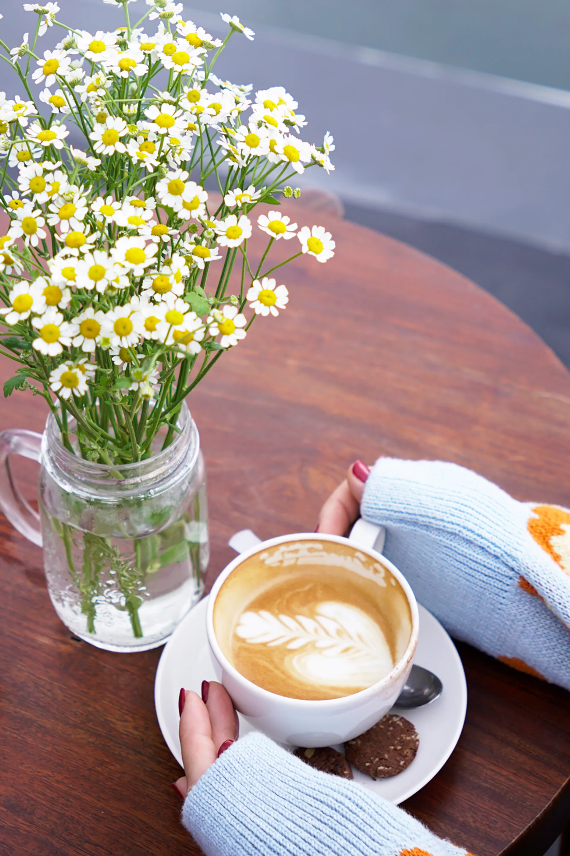 Aerial view of woman in sweater holding a cup of cafe latte on a table decorated with daisies