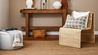 Cozy room with wicker chair, plaid pillow, wooden console, books, lamp, and decorative vases on natural fiber rug.