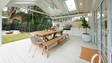 Covered outdoor patio with wooden dining table, benches, barbecue grill, and potted plants in a garden setting.