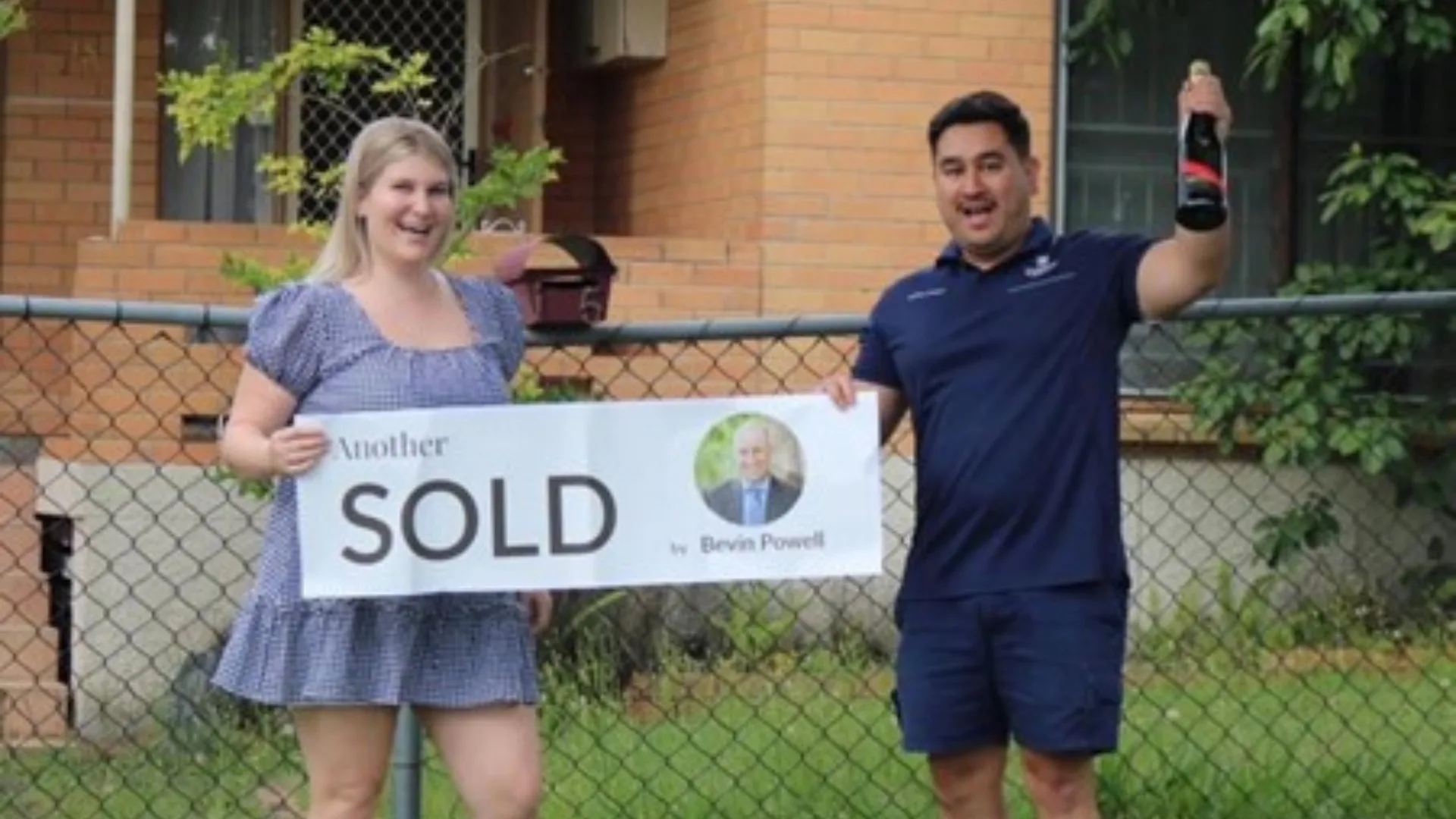 couple standing outside of brick house holding sold sign and bottle of champagne