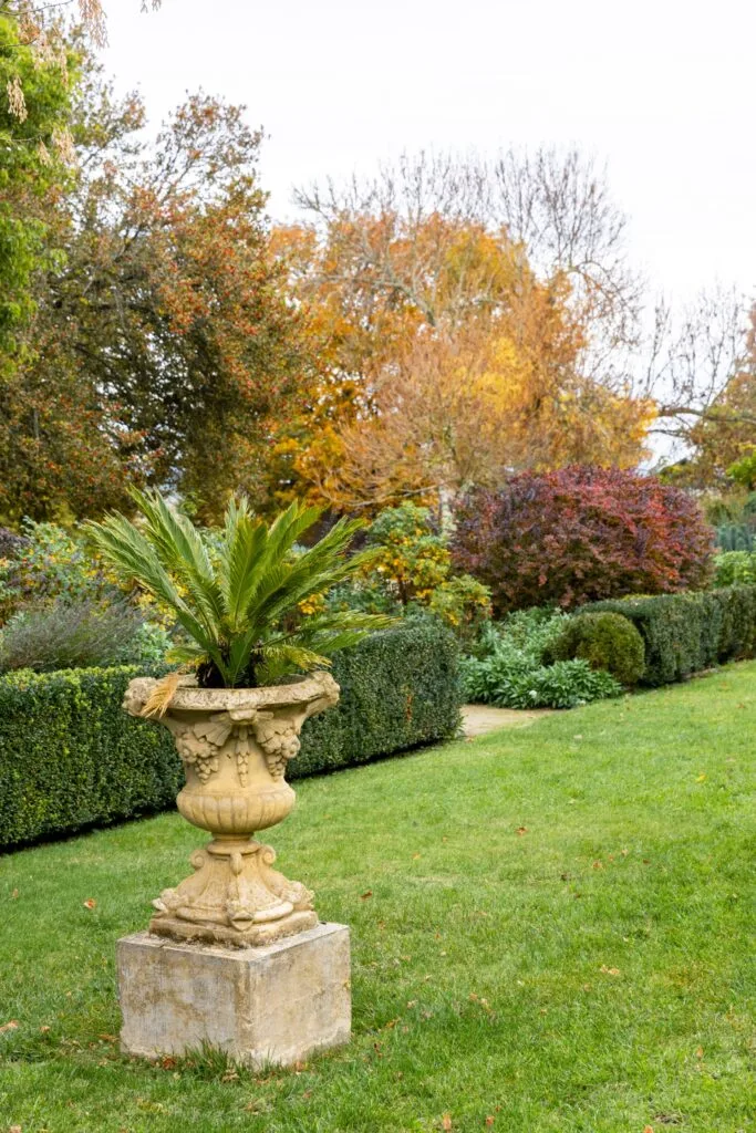 ornamental stone pot with fern in it on a long grass lawn with hedged garden