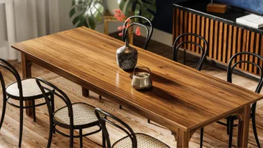 Wooden dining table with black chairs, a flower vase, and a teapot on top, in a room with a wooden floor and sideboard.