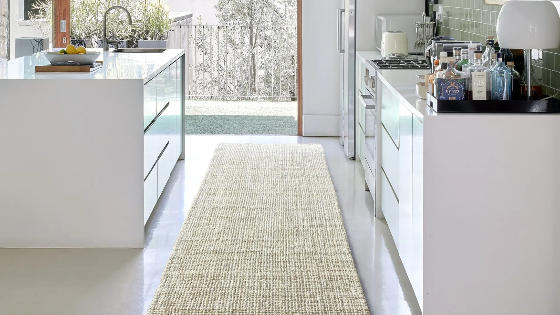 Modern kitchen with white cabinets, a central island, and a beige runner rug on polished floor, leading to open garden view.