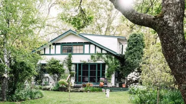 A charming, white cottage with green trim, surrounded by lush trees and a sunlit garden.