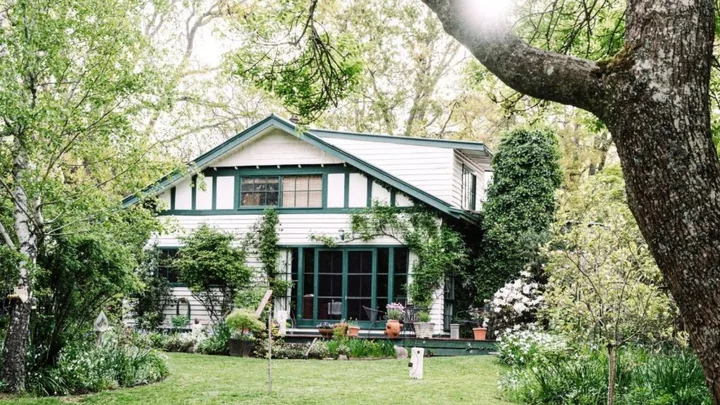 A charming, white cottage with green trim, surrounded by lush trees and a sunlit garden.