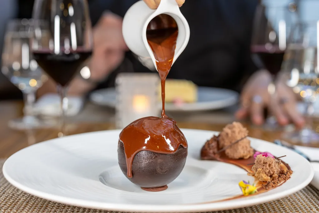 tempered chocolate ball with jug pouring melted chocolate over the top. Also on the plate are crumbled cake and a swirl of chocolate sauce
