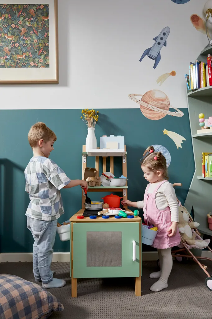 kids play kitchen with two little children standing either side of it. complete with tiny pots and pans, cups and plastic kitchen vegetables