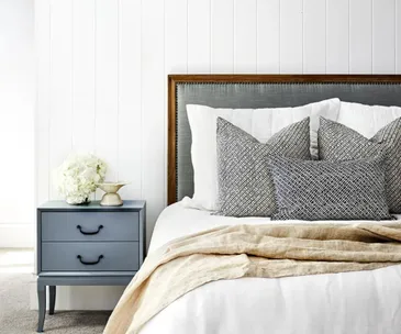 Cozy bedroom with gray headboard, layered pillows, beige throw, and blue nightstand with flowers.