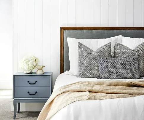 Cozy bedroom with gray headboard, layered pillows, beige throw, and blue nightstand with flowers.