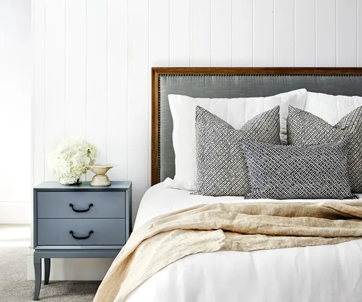 Cozy bedroom with gray headboard, layered pillows, beige throw, and blue nightstand with flowers.