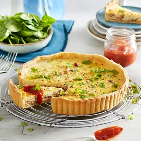 Quiche with herbs on cooling rack, slice removed. Jar of chutney, salad, and plate in background.