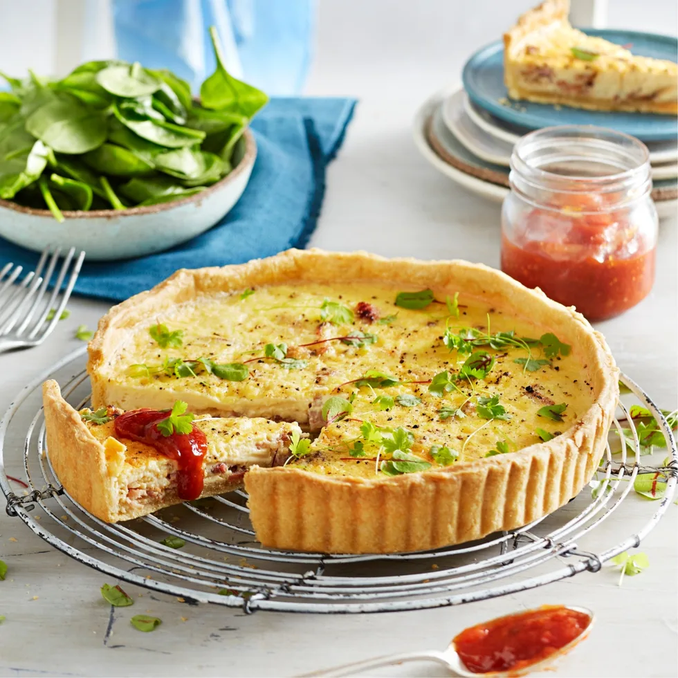 Quiche with herbs on cooling rack, slice removed. Jar of chutney, salad, and plate in background.