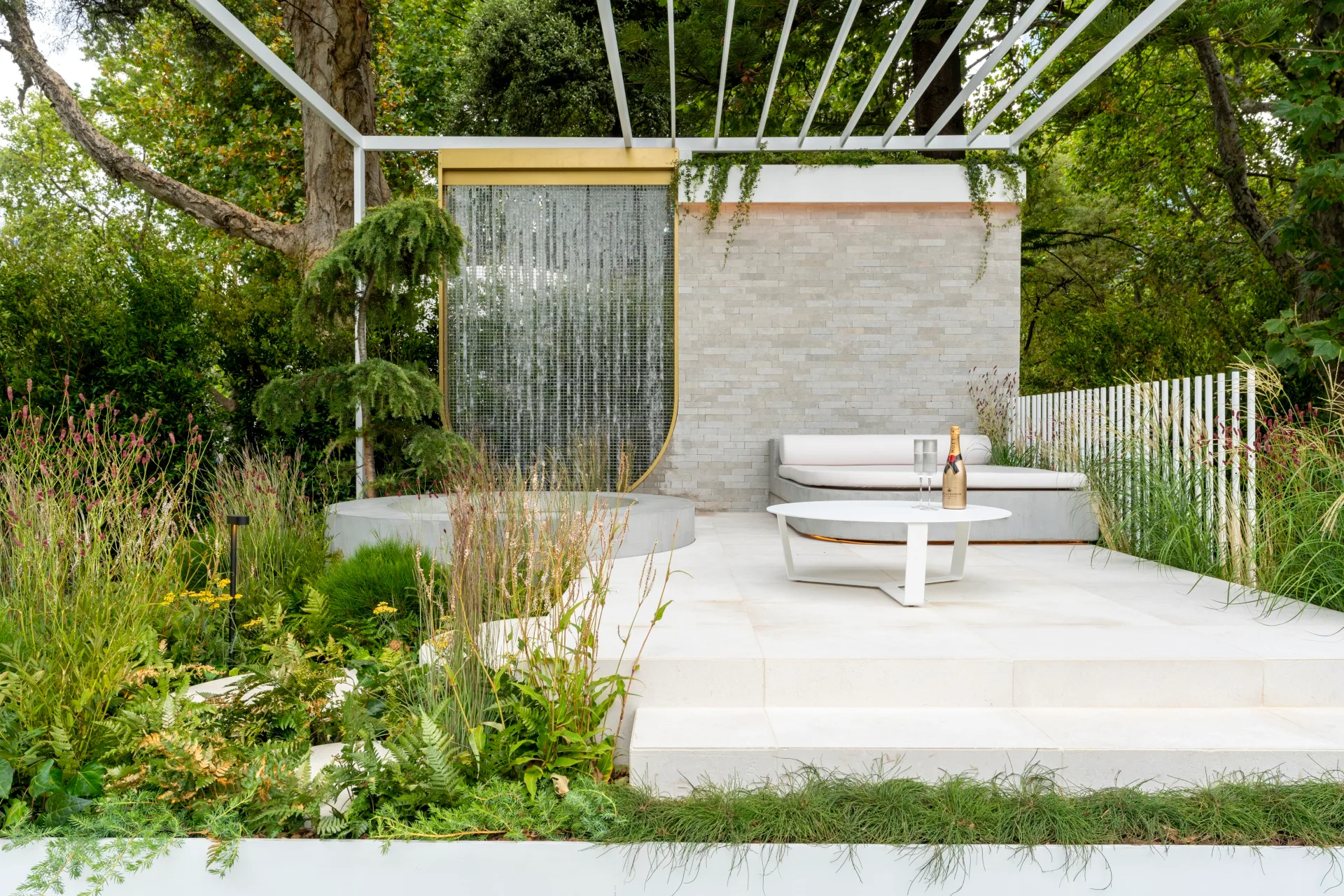 contemporary landscape garden with curtain of beaded material, step up patio and white pergola surrounding by loose, chaotic plants