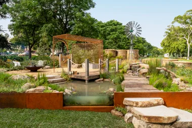 Scenic garden with a wooden path, pond, arch, fire pit, and windmill surrounded by lush greenery and trees.