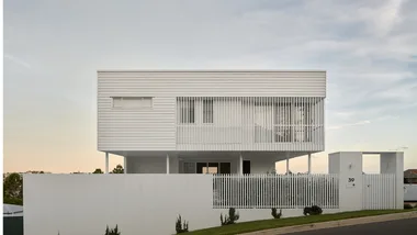 Modern white two-story house with large windows and a picket fence, set against a sunset sky.
