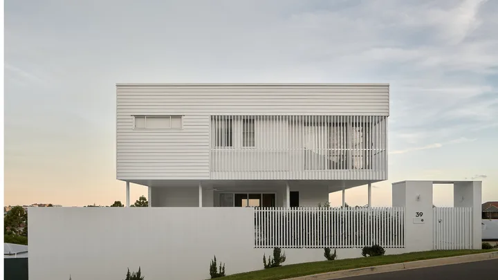 Modern white two-story house with large windows and a picket fence, set against a sunset sky.
