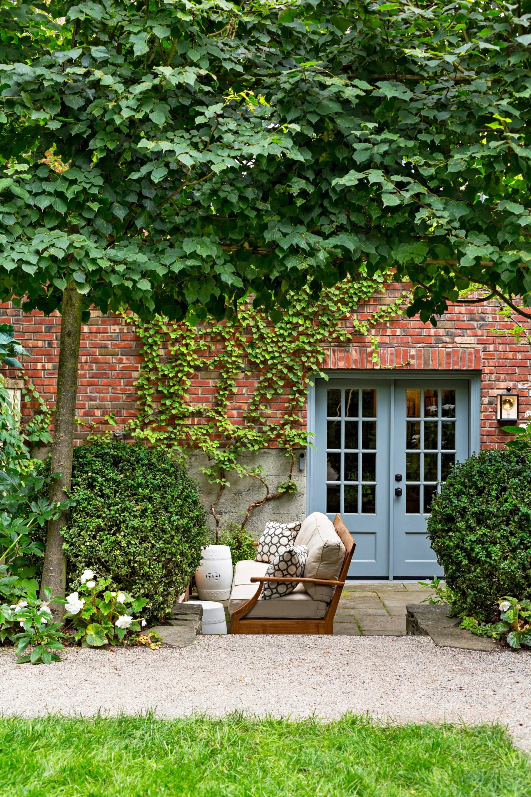 create an outdoor living space with side view of red brick house with blue french doors, outdoor seating with wooden frame and creeping ivy up the wall. In front a grass lawn and to the right a large leafy tree