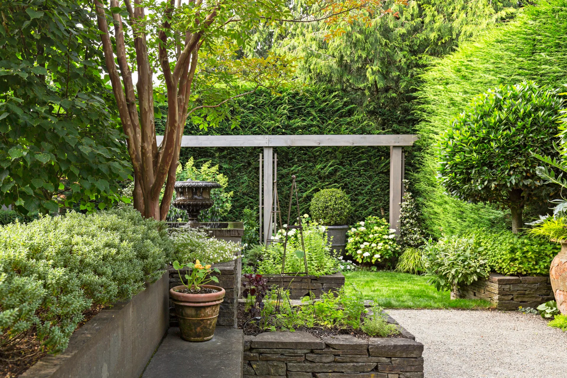 raised garden beds made of slate stone with small trellis' on them. surrounded by leafy green hedging and small trees with potted plants dotting the space.