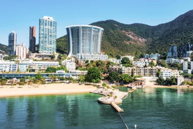 Beachfront cityscape with tall buildings and greenery, set against a background of lush hills and clear blue sky.