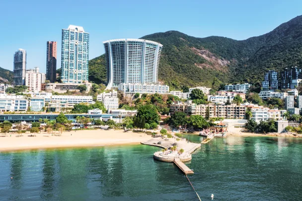 Beachfront cityscape with tall buildings and greenery, set against a background of lush hills and clear blue sky.