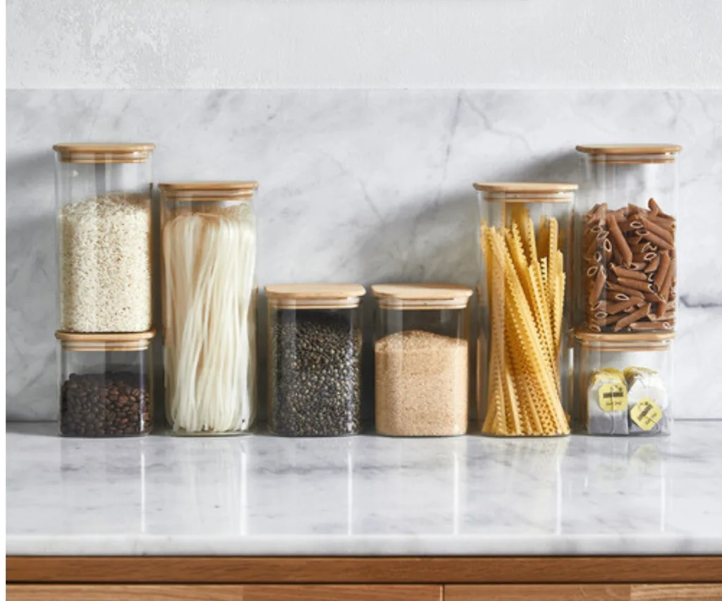 7 different sized glass containers filled with non perishables like pasta, noodles and rice