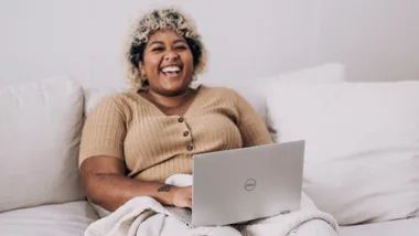 A woman sitting on a couch laughing with a Dell computer on her lap.