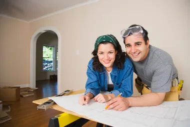 Couple smiling, leaning on a table with house plans in a room under renovation.