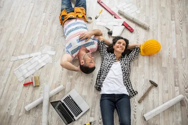Couple lying on wooden floor among tools and a laptop, smiling and looking relaxed.