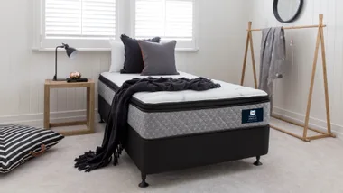 Single bed in a modern bedroom with black and grey pillows, a blanket, a wooden side table with a lamp, and a clothes rack.