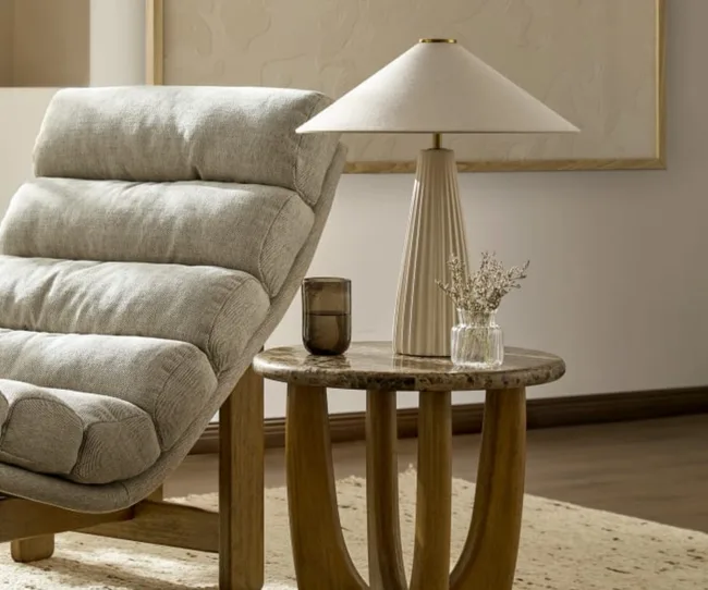 Cozy gray armchair next to round wooden table with a modern lamp, glass, and small vase with dried flowers.