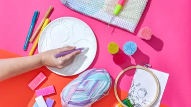 Crafting supplies: a hand using a purple pen on embossed plate, pastel yarn, markers, a hook tool, and embroidery set.