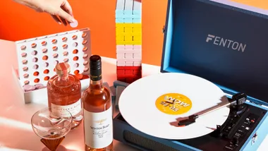 Hand placing token in game next to bottles and glasses, colorful block tower, and blue Fenton record player with white vinyl.