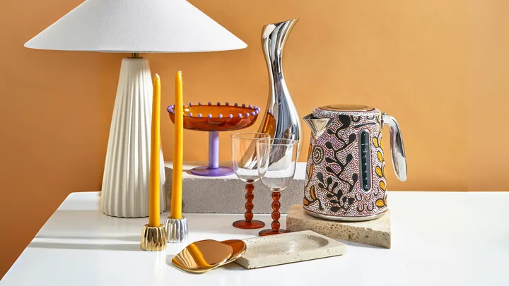 Home decor gifts arranged on a white table including a lamp, brass candle holders, coasters and more.