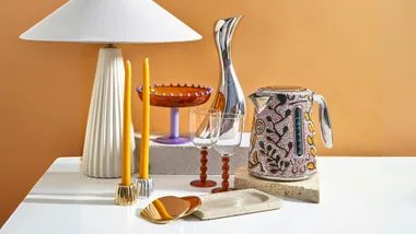 Home decor gifts arranged on a white table including a lamp, brass candle holders, coasters and more.