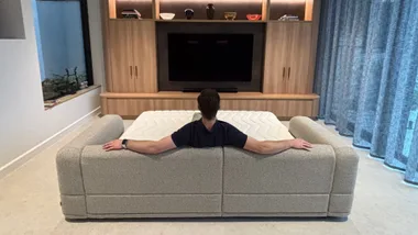 Man sitting on sofa bed in a living room, facing a large TV and bookshelf, arms resting on backrest.