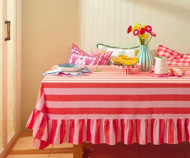 Striped pink and red tablecloth with colorful cushions, a vase of flowers, and fruit decoratively arranged on the table.