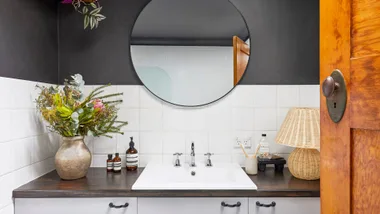 Bathroom vanity with round mirror, vase with flowers, bottles, and woven lamp on dark countertop.