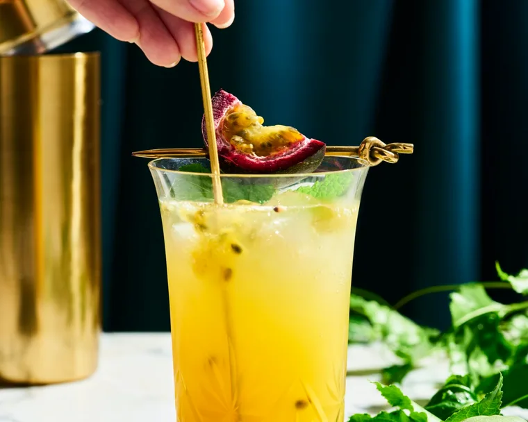 a tall glass with yellow liquid in it with passionfruit seeds swirling in the liquid. This is on a marble table top with a dark navy background. You can find a cut slice of passionfruit next to the glass on the right side, as well as, passionfruit vine behind the glass.