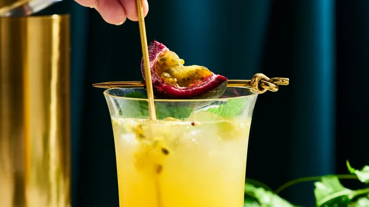 a tall glass with yellow liquid in it with passionfruit seeds swirling in the liquid. This is on a marble table top with a dark navy background. You can find a cut slice of passionfruit next to the glass on the right side, as well as, passionfruit vine behind the glass.