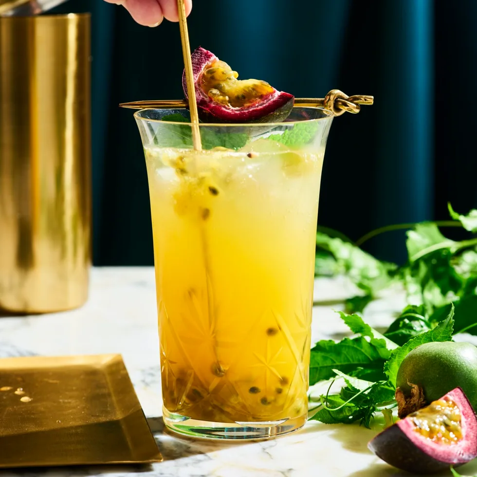 a tall glass with yellow liquid in it with passionfruit seeds swirling in the liquid. This is on a marble table top with a dark navy background. You can find a cut slice of passionfruit next to the glass on the right side, as well as, passionfruit vine behind the glass.