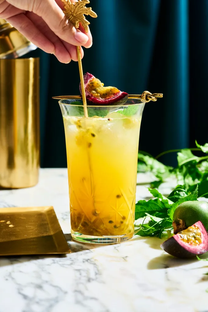 a tall glass with yellow liquid in it with passionfruit seeds swirling in the liquid. This is on a marble table top with a dark navy background. You can find a cut slice of passionfruit next to the glass on the right side, as well as, passionfruit vine behind the glass.