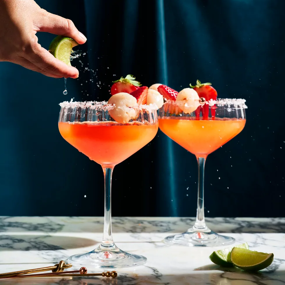 cocktail glass with strawberry liquid in it, on the right side of each glass they have put a lychee for decoration. This is on a marble board sitting in front of a dark blue background
