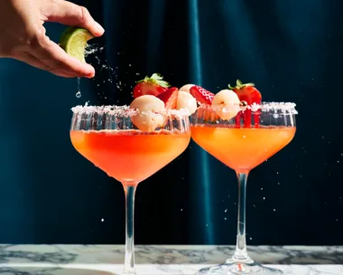 cocktail glass with strawberry liquid in it, on the right side of each glass they have put a lychee for decoration. This is on a marble board sitting in front of a dark blue background