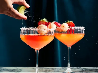cocktail glass with strawberry liquid in it, on the right side of each glass they have put a lychee for decoration. This is on a marble board sitting in front of a dark blue background