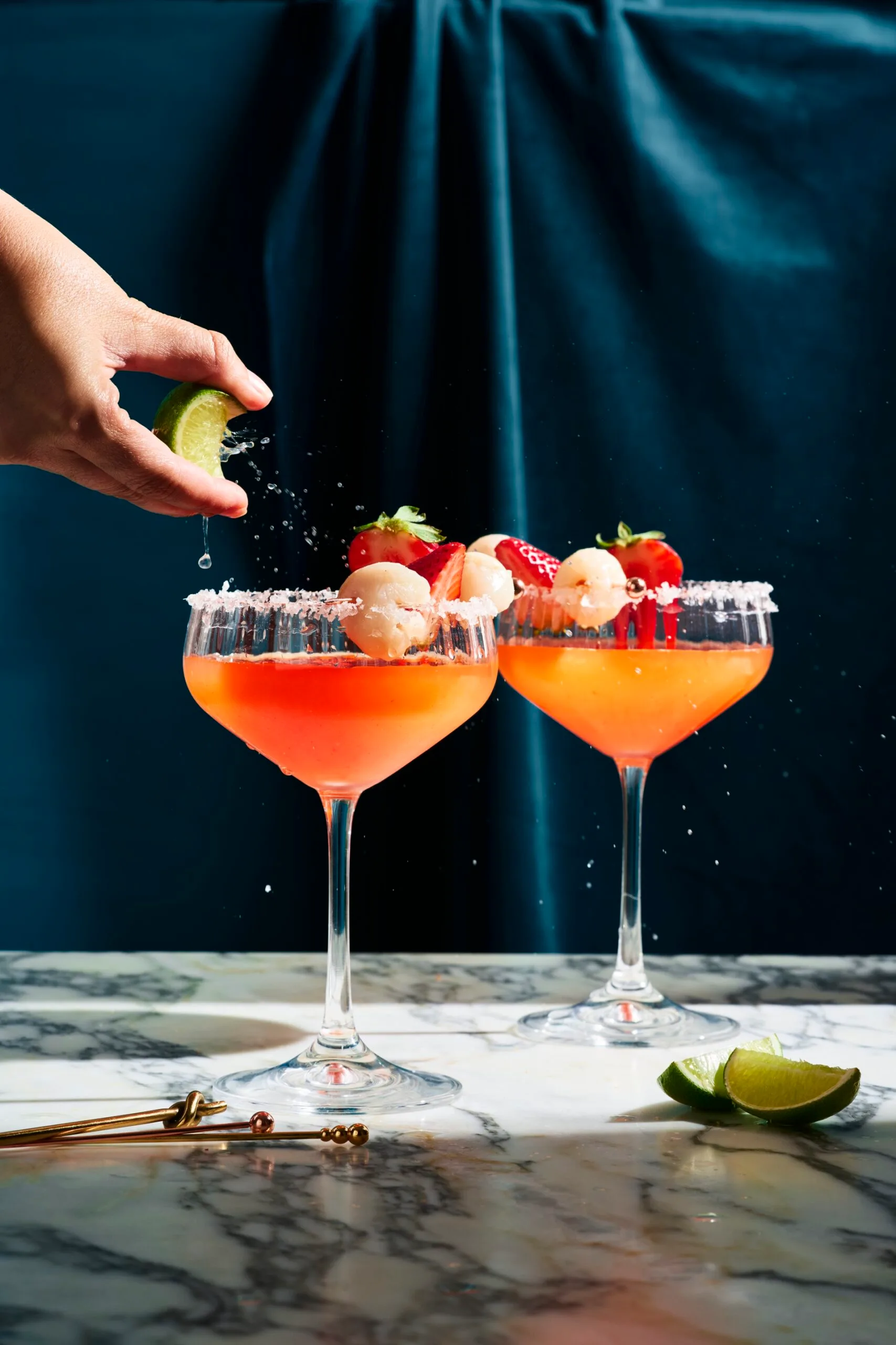 cocktail glass with strawberry liquid in it, on the right side of each glass they have put a lychee for decoration. This is on a marble board sitting in front of a dark blue background