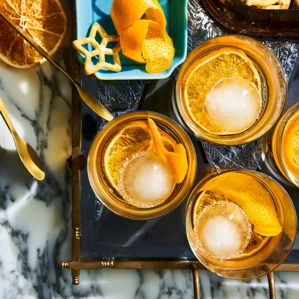 A birds eye view looking at 4 old fashioned cocktails in a short tumbler glass, they have a twisted orange peel in them as a garnish. They are placed on a marble table top with a light blue rectangular plate to the left which has more garnishes on it