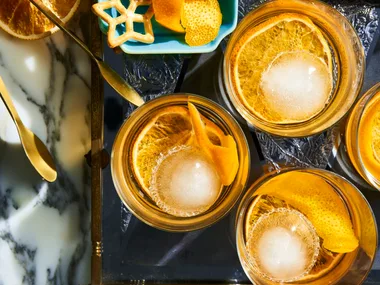A birds eye view looking at 4 old fashioned cocktails in a short tumbler glass, they have a twisted orange peel in them as a garnish. They are placed on a marble table top with a light blue rectangular plate to the left which has more garnishes on it