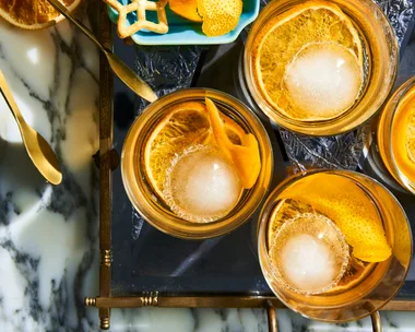 A birds eye view looking at 4 old fashioned cocktails in a short tumbler glass, they have a twisted orange peel in them as a garnish. They are placed on a marble table top with a light blue rectangular plate to the left which has more garnishes on it