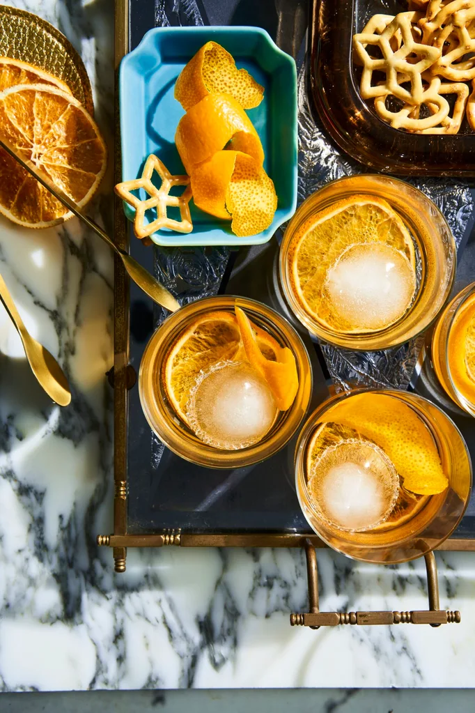 A birds eye view looking at 4 old fashioned cocktails in a short tumbler glass, they have a twisted orange peel in them as a garnish. They are placed on a marble table top with a light blue rectangular plate to the left which has more garnishes on it