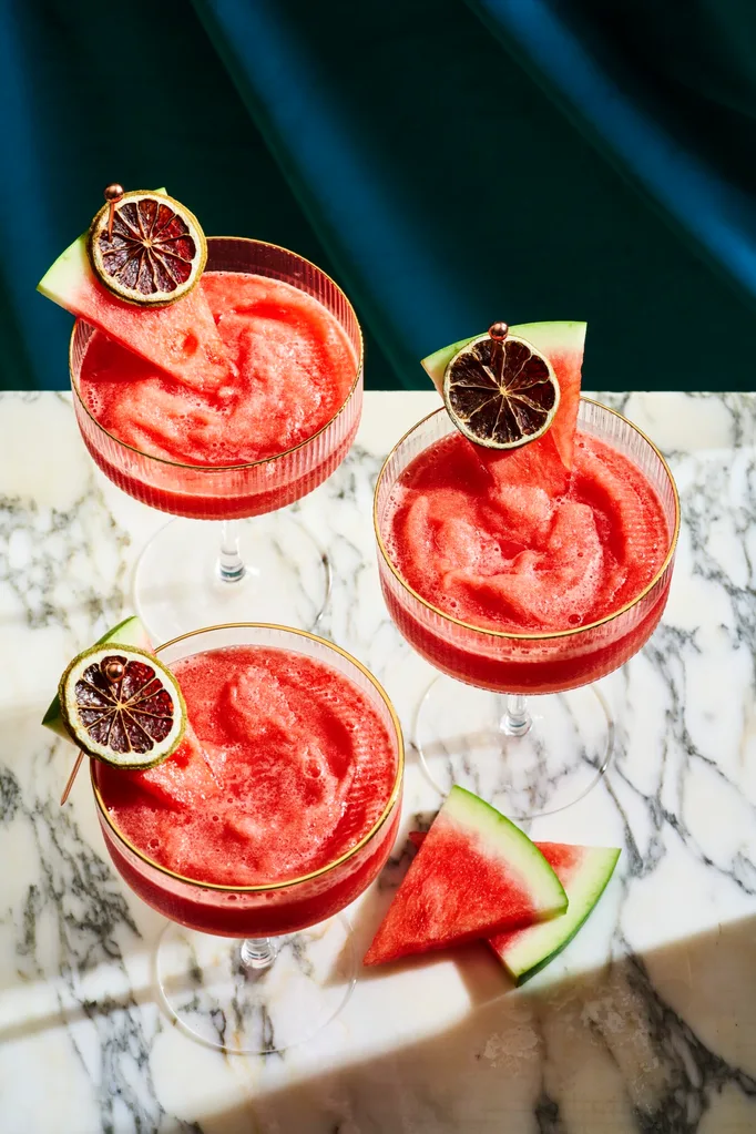 3 watermelon daiquiri's on a marble board, with a wedge of watermelon and a dried lime ontop of the cocktail as a garnish